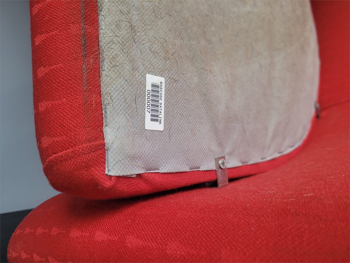 A small white asset label reading BARCODE DATALINK above a barcode and the number 000007, on the underside backing board of a red office chair.