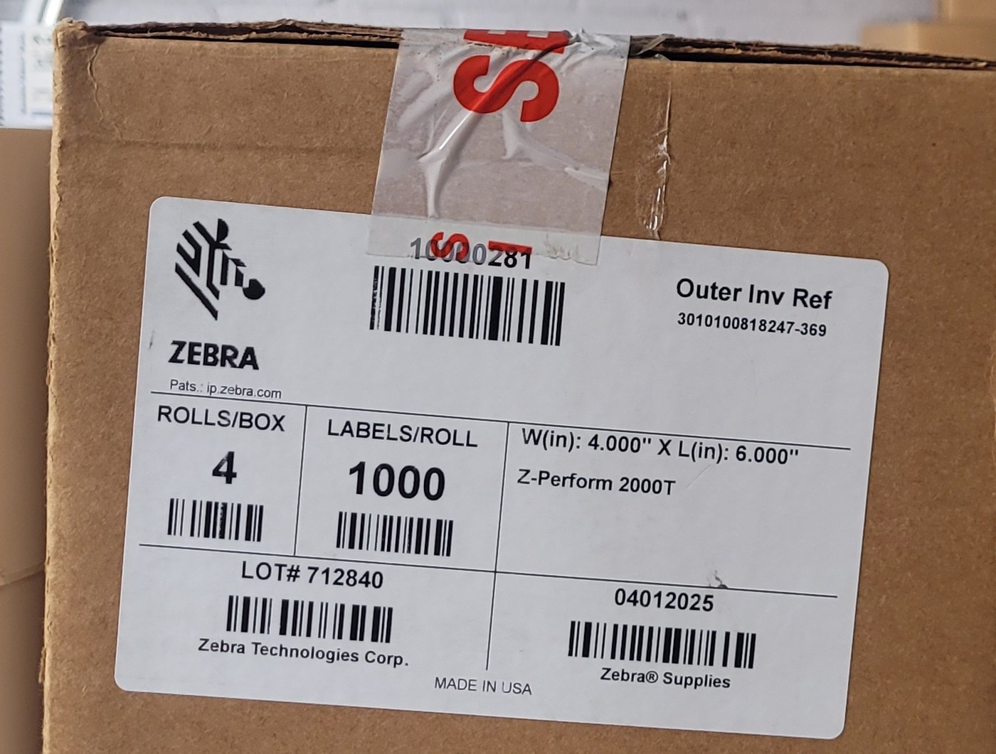 Close view of the top of a Zebra label carton showing red and white SECURITY SEAL tape across the flap, above a carton label reading Zebra Technologies Corp and MADE IN USA.