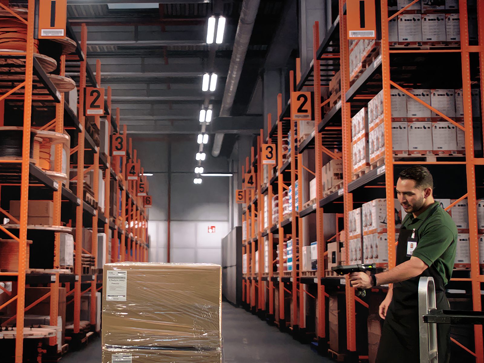 A long warehouse aisle with orange racking, numbered bay labels running away
                  into the distance, and an operator at the far end scanning with a gun-grip
                  Zebra device beside a wrapped pallet.