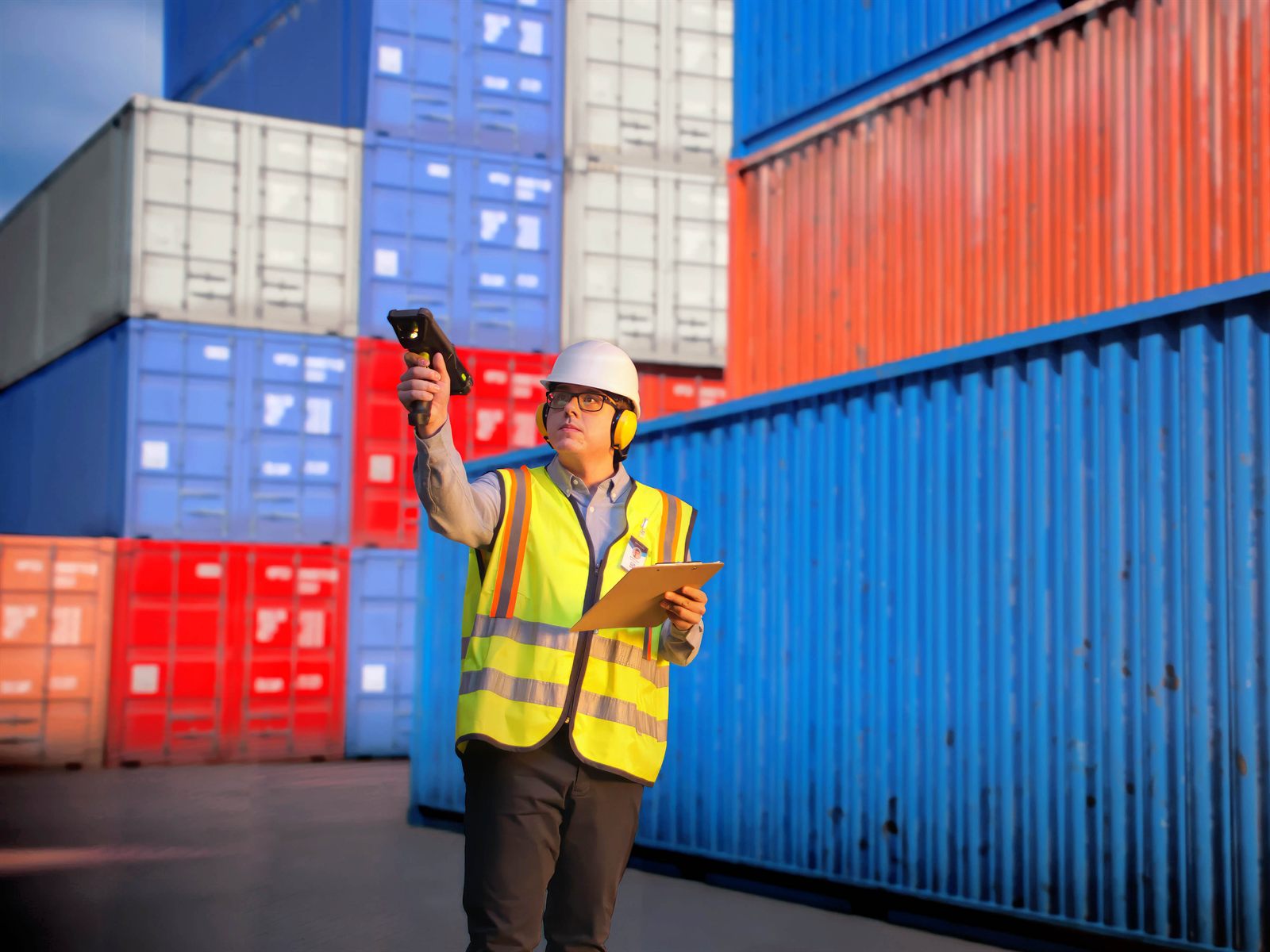 A worker in a hard hat and hi-vis vest in a container yard, aiming a gun-grip
                    Zebra MC9400 upward at a label on a stacked shipping container while holding a
                    clipboard in the other hand.