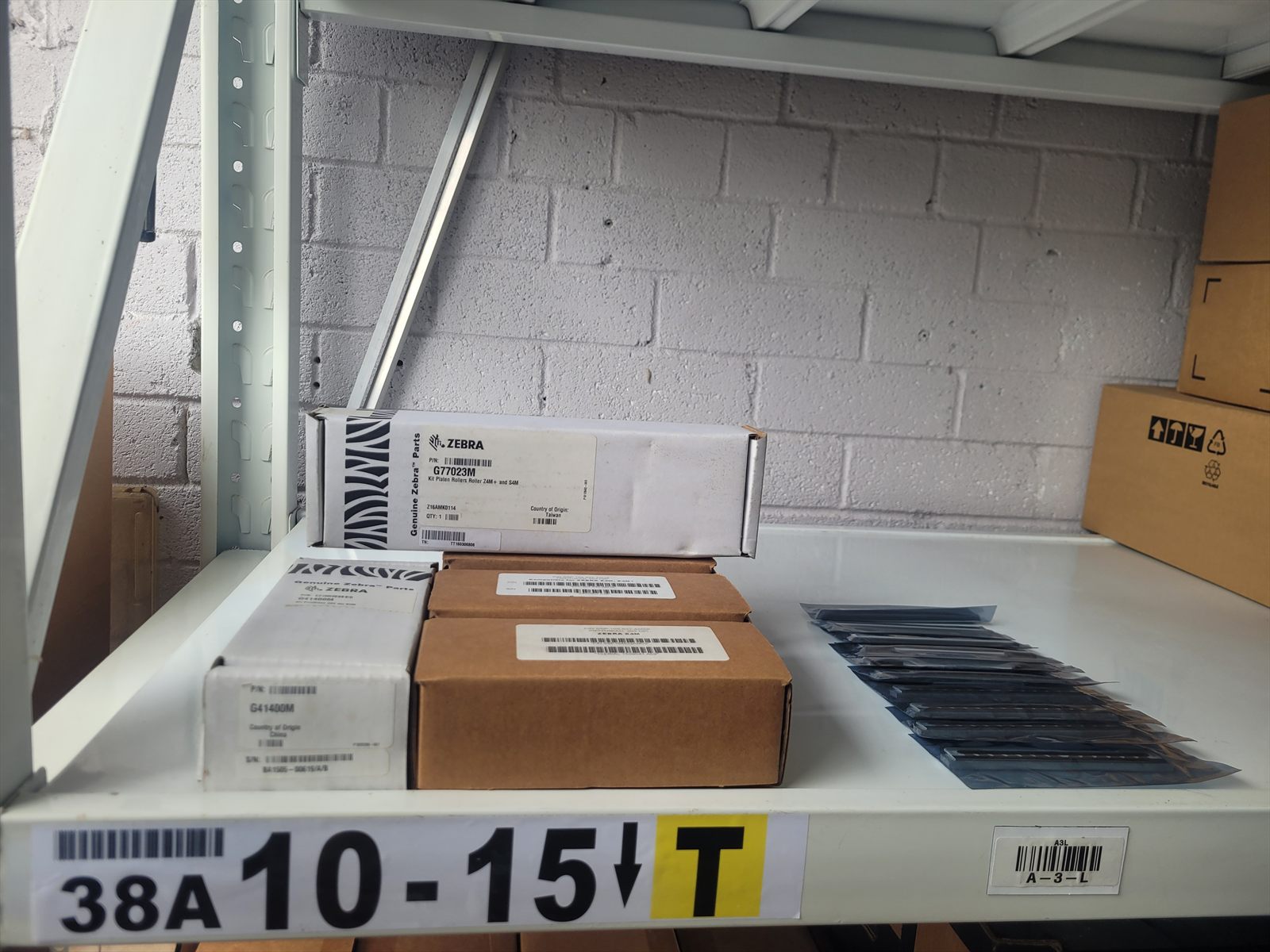 A steel warehouse shelf holding boxed Zebra printhead and platen roller kits, with a row of bare printheads in anti-static bags beside them. A barcoded bin label reading 38A 10-15 is fixed to the shelf edge.