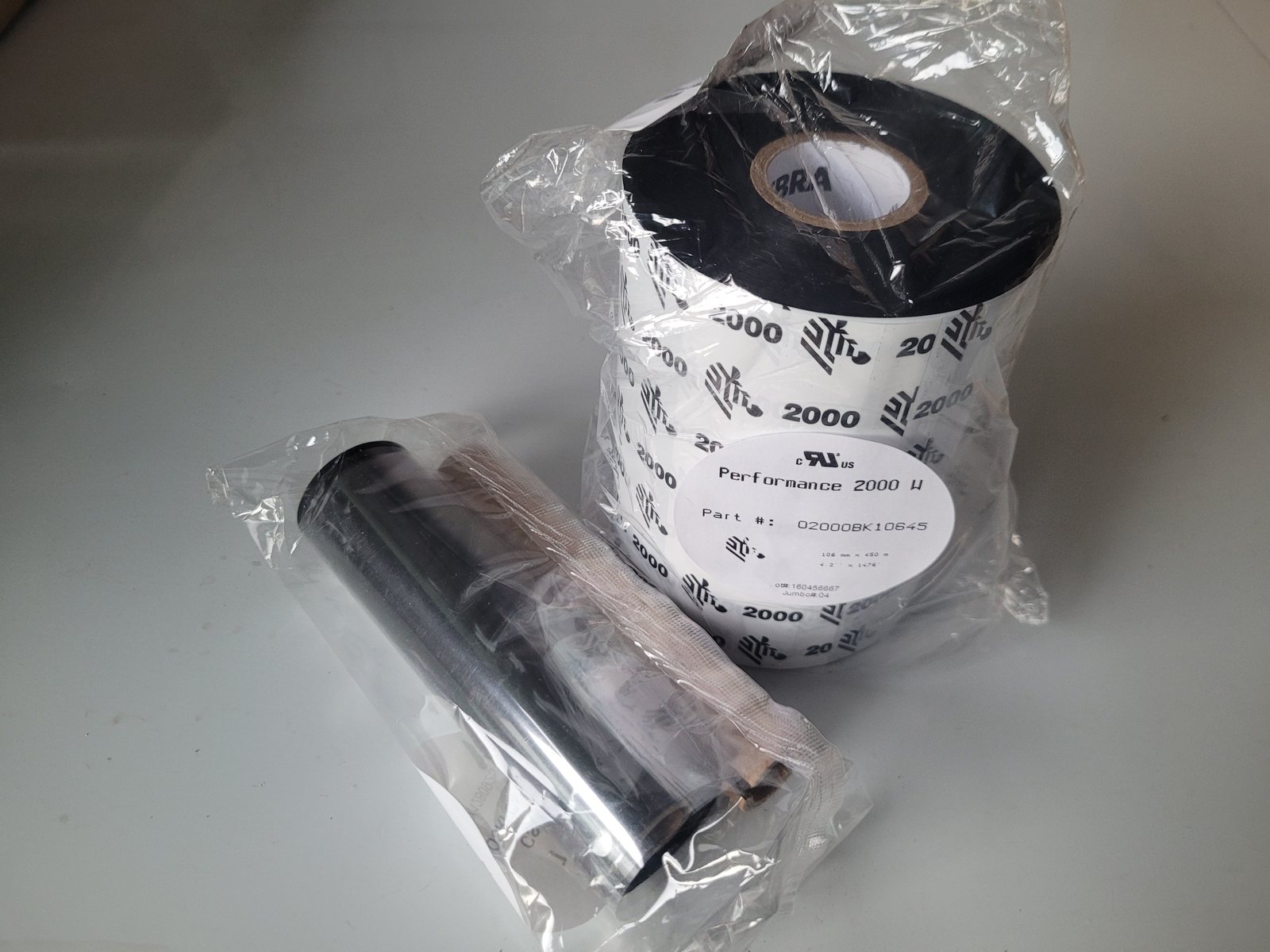 An industrial thermal transfer ribbon beside a desktop one. The industrial
                  roll is several times the diameter and sits on a wide core; the desktop
                  ribbon is a slim roll on a much smaller core.