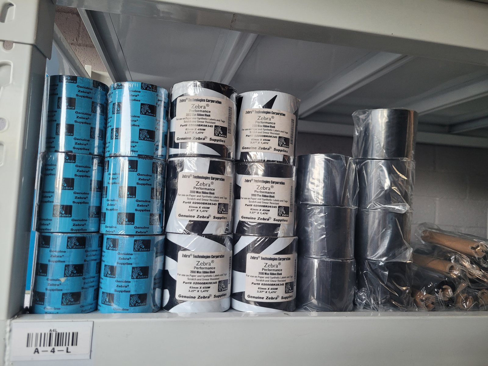 A shelf of Zebra thermal transfer ribbons in their wrappers, the front boxes
                  labelled Zebra Performance 2000 Wax Ribbon Black, 83mm by 450M, with a
                  barcoded bin label reading A-4-L on the shelf edge