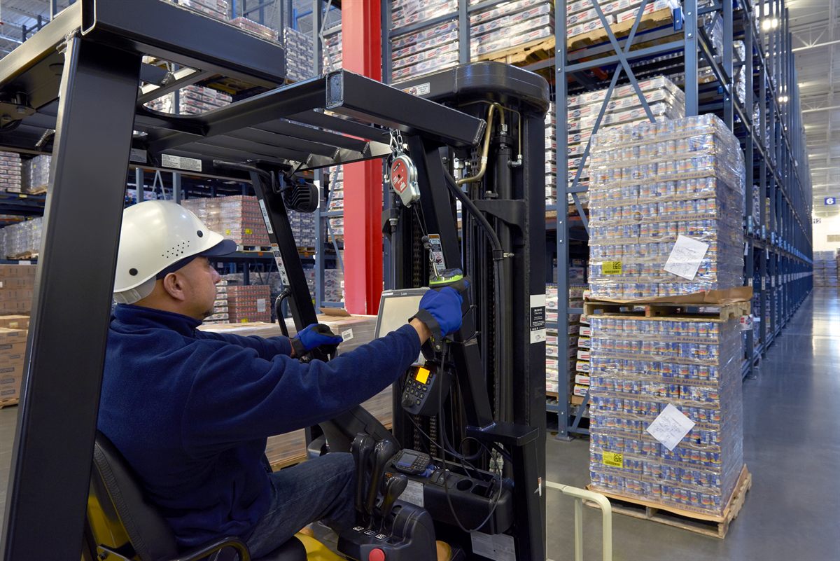 A forklift operator in a distribution centre reaching for a vehicle-mounted terminal, a rugged green and black Zebra scanner in his gloved hand, high racking and wrapped pallets around him.
